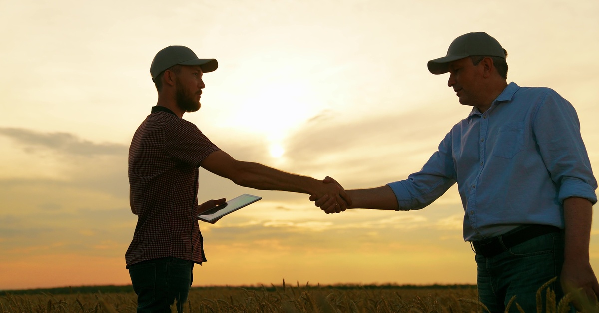 Two men in hats shaking hands while standing in some fields of wheat. The sun is setting behind the men.