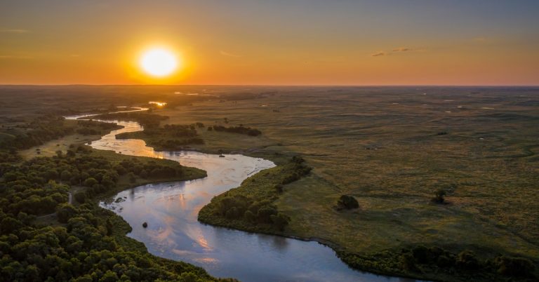 A river cutting through the land. There is farmland on one side and a forest on the other. The sun is setting on the river.