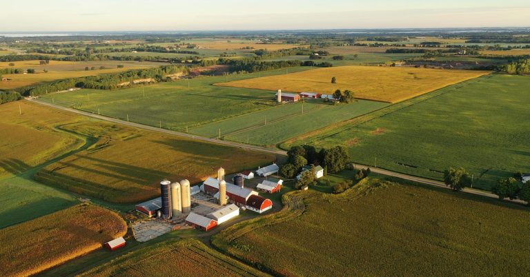 An overview of some farmland. There is a large farm with multiple barns and silos in the middle of the land.
