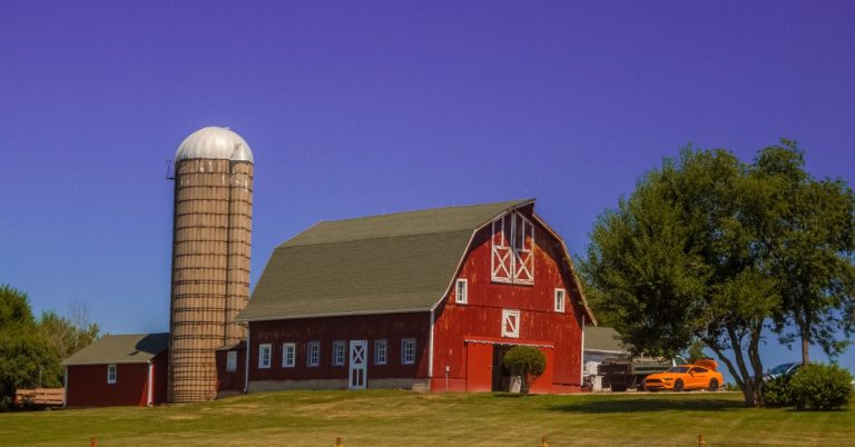 A large red barn with a silo and small shed behind it. There are also some vehicles parked off to the side.