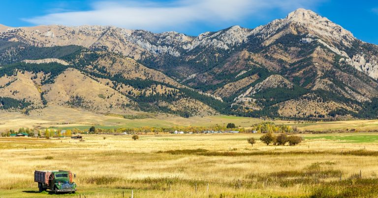 A section of farmland that leads up to a large mountain range. There is a vintage truck parked in one of the fields.