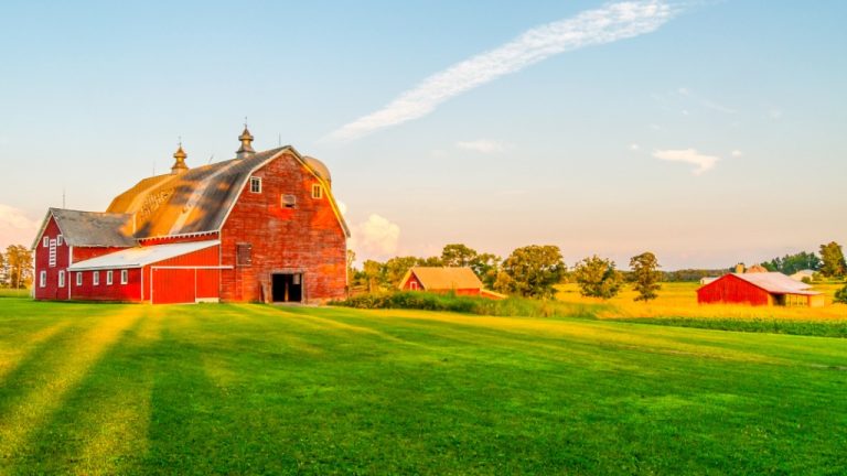 A farmstead at sunset with grassy fields, trees, a red barn, and soft clouds across a wide rural sky.
