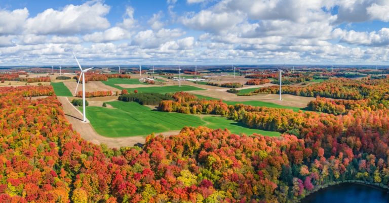 An autumn farm landscape with wind turbines, crop fields, and groves of trees. The sky is filled with puffy white clouds.
