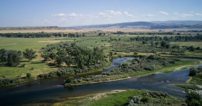 A winding river curving through green fields and trees, with pale hills and blue sky stretching beyond the valley.
