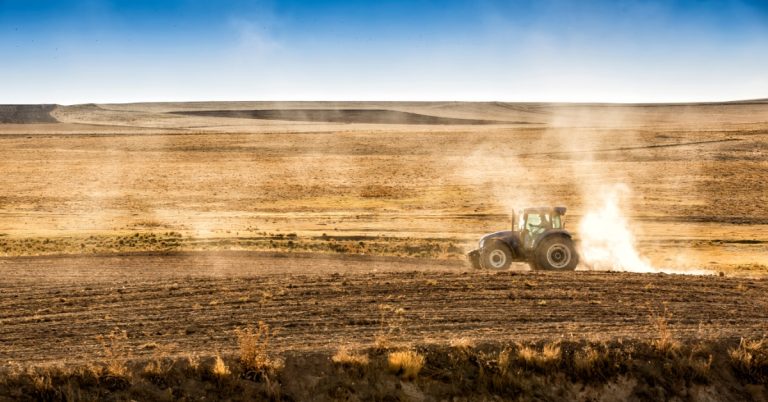 A tractor driving across a dry brown field while kicking dust up behind it. The field stretches far out into the distance.