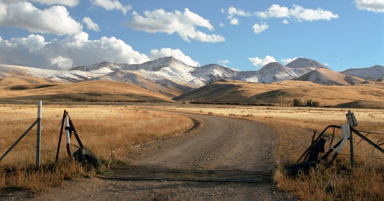 A ranch entrance gate standing beside a gravel road, with snowy mountains and open sky across the wide landscape.