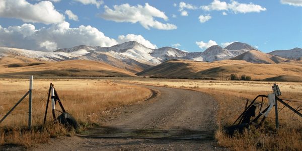 A ranch entrance gate standing beside a gravel road, with snowy mountains and open sky across the wide landscape.
